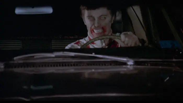 Undead soldier with blood on his face drives a car with a menacing grin in a scene from Dead of Night (1974).
