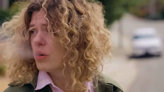Woman with curly hair exhales smoke while looking over her shoulder on a suburban street