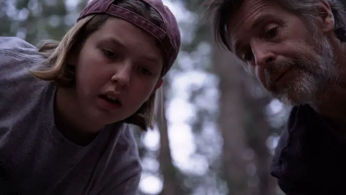 Teenage girl in a backward cap and her bearded father look down intently in a forest setting.