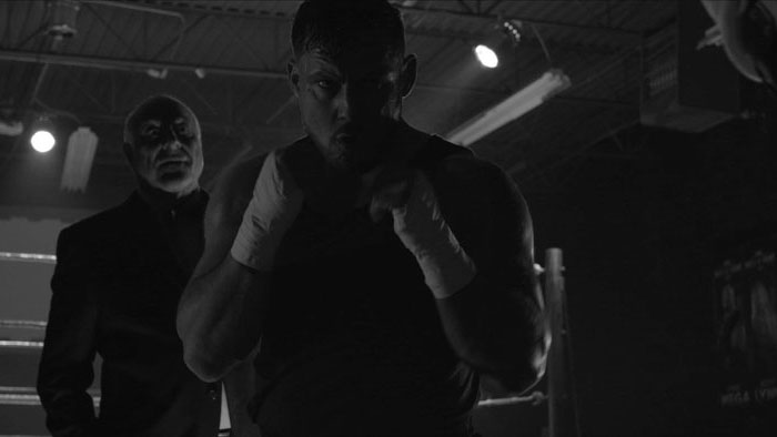 A boxer stands in a dimly lit gym with his fists raised while an older man watches behind him.