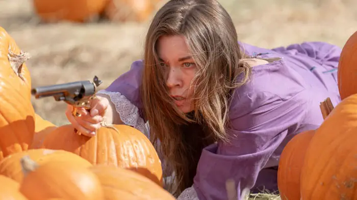 A woman in a purple dress aims a gun while hiding among pumpkins.