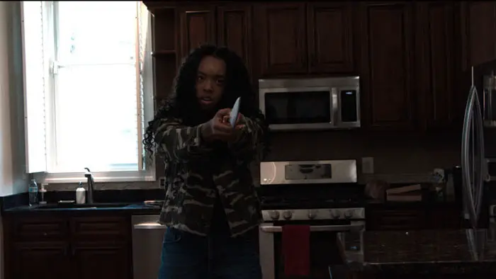 A woman in a camouflage jacket points a knife in a tense kitchen scene from The Final Podcast.