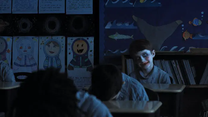 A child with smeared makeup smiles eerily in a dark classroom while other students have their heads down.