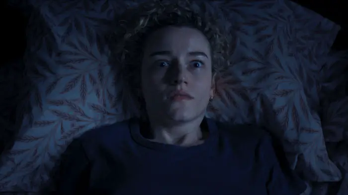 Julia Garner stares wide-eyed in bed, illuminated in blue light.