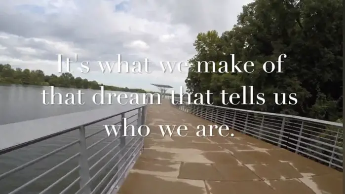 Text overlay on a riverside walkway in Charming Blue: “It’s what we make of that dream that tells us who we are.” 