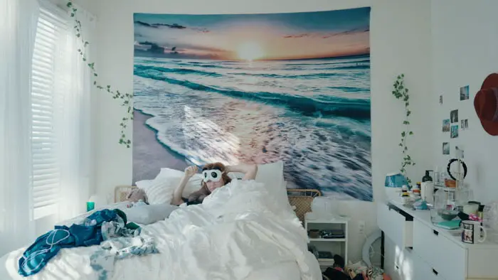 Woman lying in bed with owl eye mask under a beach sunset wall tapestry.