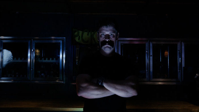 A shadowy man with a mustache stands in a dimly lit bar scene from Pickle Vision.