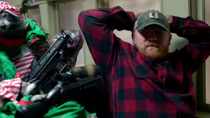 A man in a red plaid shirt held hostage by a raccoon dressed as an elf and armed with a rifle in Killer Raccoons 2: Dark Christmas in the Dark.