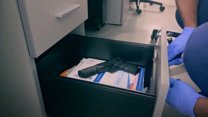 A gloved hand opens a desk drawer revealing a handgun inside a medical office.