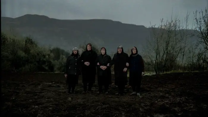 Five women stand solemnly in a barren field under a dark sky in Sun Never Again. 