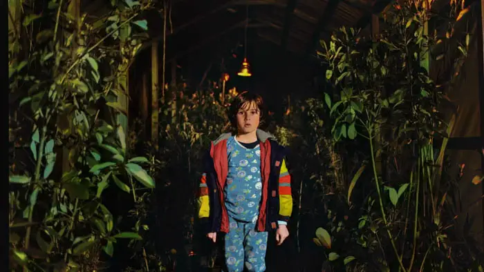 A young boy stands in a greenhouse filled with lush plants in Sun Never Again.
