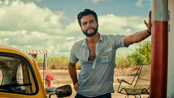 Wagner Moura in The Secret Agent (2025) stands by a yellow car at a rural gas station in Recife, Brazil.