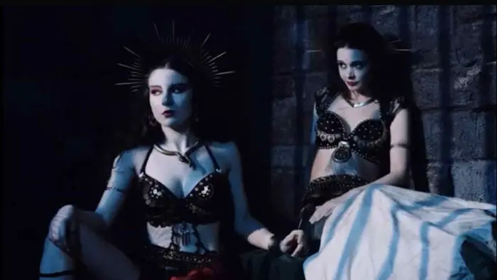 Two dark-clad sorceresses sit in a stone chamber under blue light.