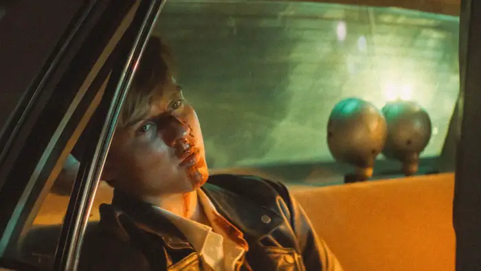 Maxwell Jenkins as Simon sitting bloodied in the back of a police car in This Too Shall Pass (2025).