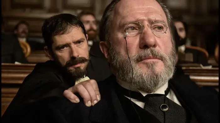 Two men in period attire sit in a crowded courtroom in An Officer and a Spy