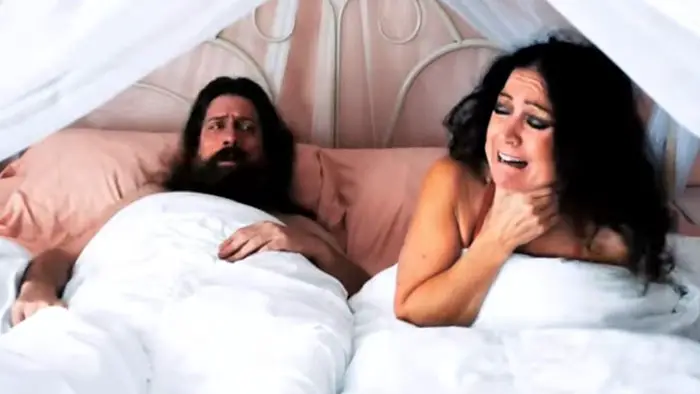 A terrified woman reacts in bed beside a bearded man in Jacker 3: Road to Hell 