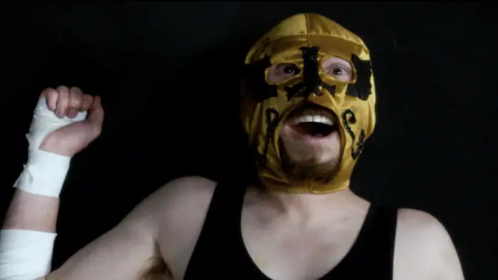 Little Lucha lies on the mat in his gold mask, laughing with his arm raised. 