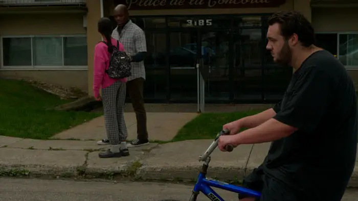 A student rides a blue bike past a girl talking with another student in front of an apartment building.