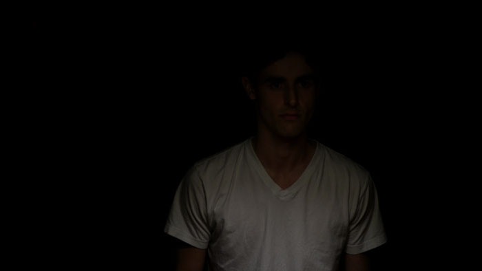 A young man partially lit in a dark room wearing a white T-shirt. 