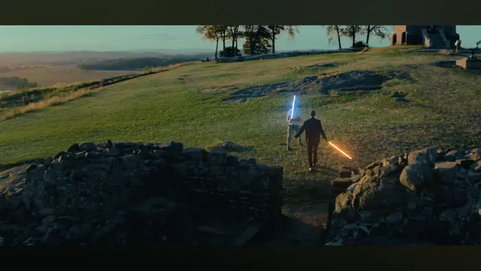 Two men duel with glowing sabers on a grassy hill overlooking the ruins of Slottsfjellet in Norway. 