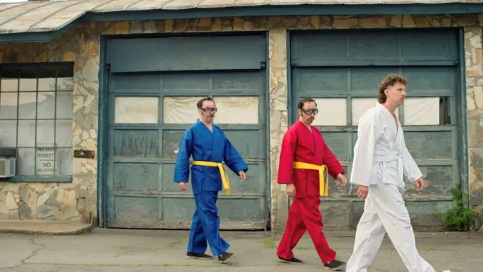 Jon Heder walks in a white karate gi alongside two men in blue and red gis outside an old garage in Tapawingo.