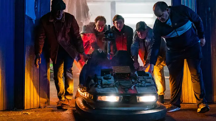 Group of characters pushing a glowing, makeshift time-travel bumper car out of a garage at night.