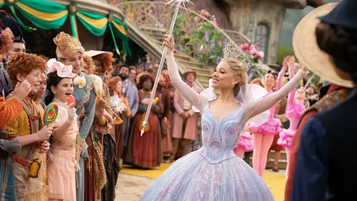 Ariana Grande as Glinda spreads her arms in a sparkling gown while greeting cheering citizens in Wicked: For Good.