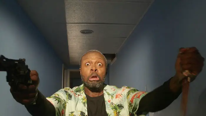 Actor in a hallway holding a gun and knife with a shocked expression in 1 Hour to Kill.