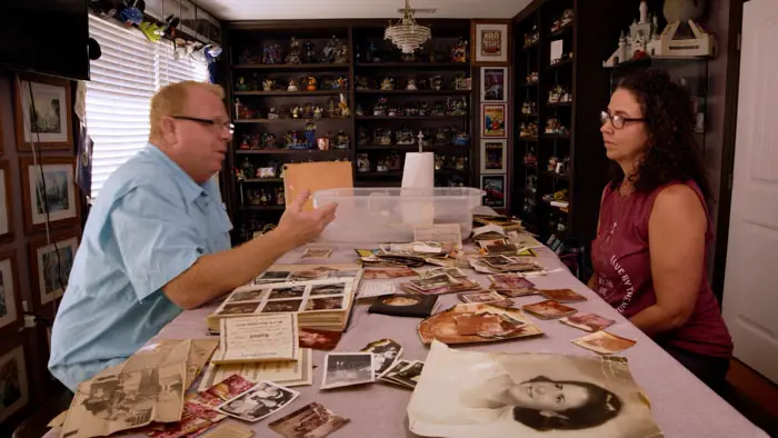 Michael Lester and Angela Lester sit at a table covered in family photos and documents in After the Devil is Dead.
