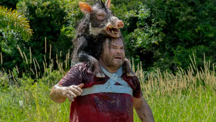 Doug McCallister staggers through tall grass with Boris the Pig clinging to his head in Anaconda (2025).
