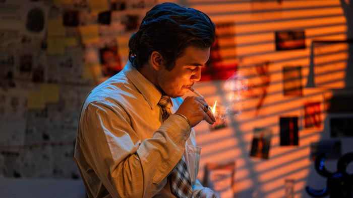 Frank smokes a cigarette in a dimly lit interrogation office with case files behind him.