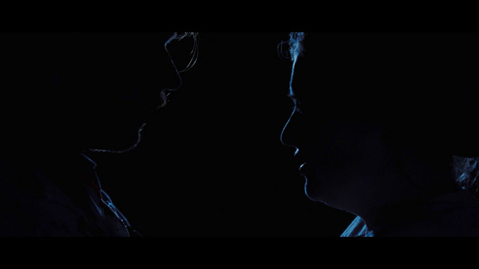 Two figures in profile face each other in near-darkness in a night still from Beyond the Pasture (2025). 