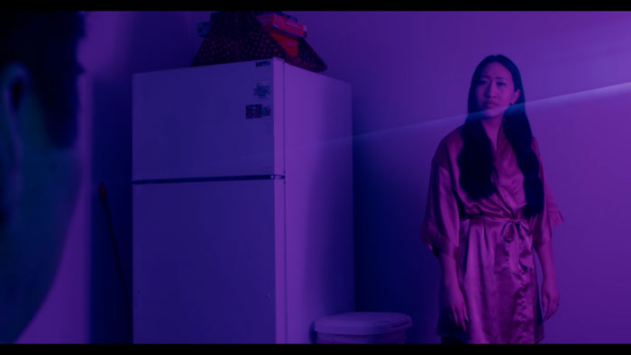 A woman stands in a purple-lit kitchen beside a refrigerator in Cho: A Tale of Voodoo (2025).