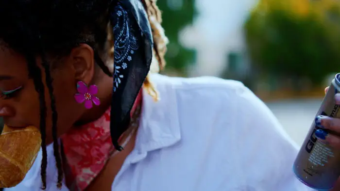 A woman wearing a bandana holds a spray can while eating bread, with her face partially obscured.