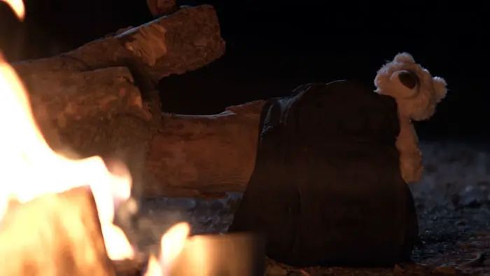 A backpack with a teddy bear peeking out sits beside a campfire in the short film Missing (2025).
