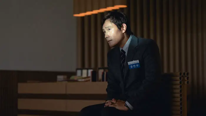 Film still from No Other Choice (2025) of Man-su (Lee Byung-hun) sitting in a suit under warm lights, wearing a name tag during a tense meeting.