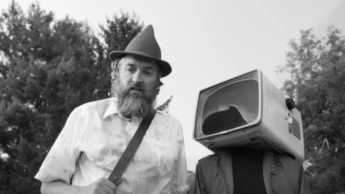 A bearded man stands beside a figure with a TV for a head in a black-and-white still from OBEX (2025). 
