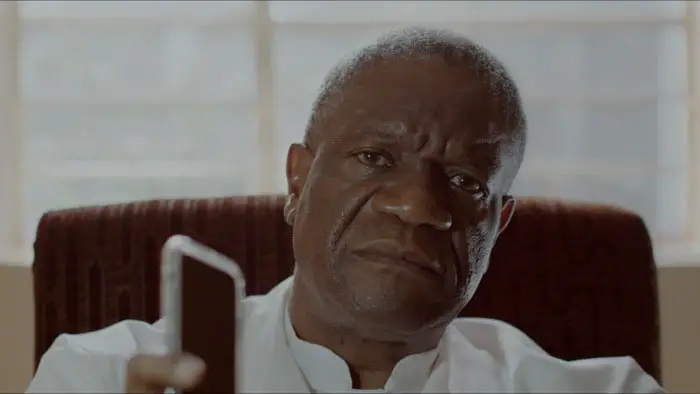 Dr. Denis Mukwege sits in his office holding a phone, looking directly ahead with a solemn expression.
