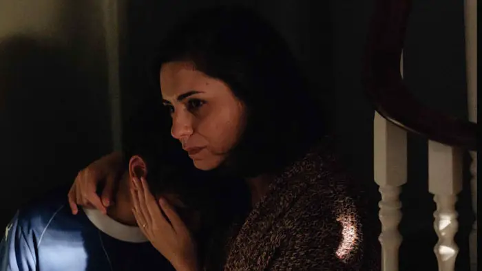 A woman sits on a staircase comforting a child in a dark, emotional moment from The Cost of Heaven (2025).