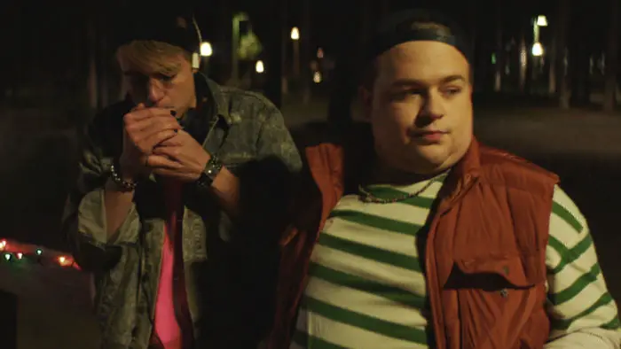 Two teens stand outside at night under warm streetlights in The Party (2026).
