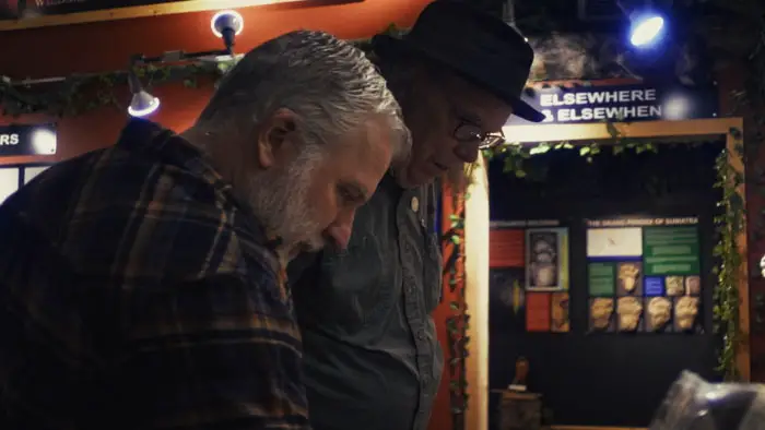 Two researchers review materials indoors while discussing the Ape Canyon Bigfoot legend in The Siege of Ape Canyon (2025).
