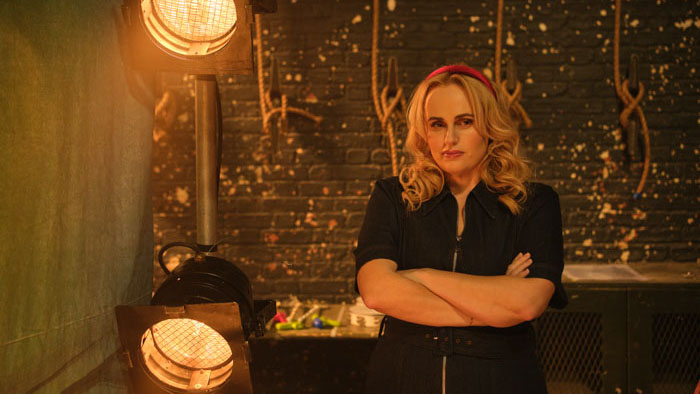 Blonde woman with arms crossed standing near stage lights in a backstage area. 