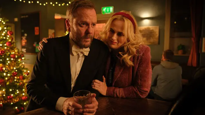 Man in tux at a pub bar as a blonde woman leans on him, Christmas tree lights in background. 
