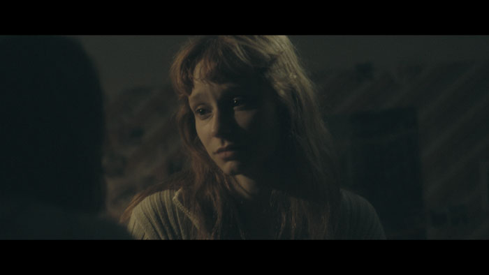 Sara Klimoska as Lena looks emotionally distressed in a dimly lit room in Unshattered.