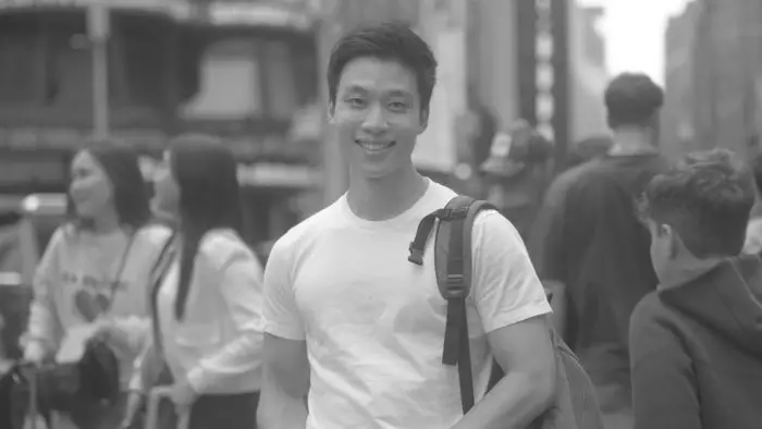 Black-and-white still from Walter Kim’s Banana Split showing Peter Lee (Min-Gu) smiling in a busy New York City street. 