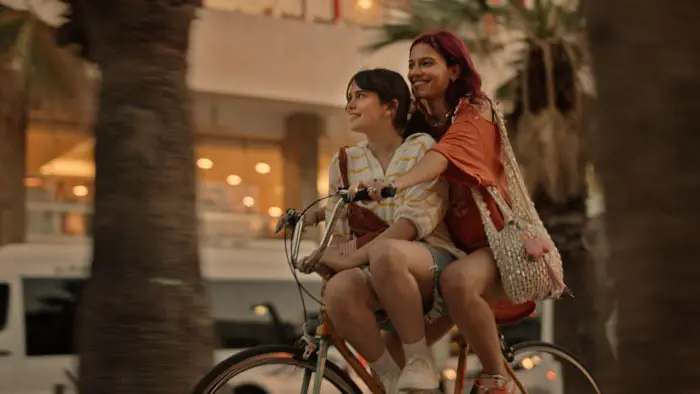 Two young women ride a bicycle together at sunset in Come Closer (2025).