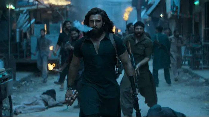 A man strides through a smoky street holding a rifle as chaos erupts around him in Dhurandhar (2025).