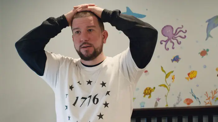 Man wearing a “1776” shirt stands indoors with his hands on his head in a scene from Homegrown (2025). 