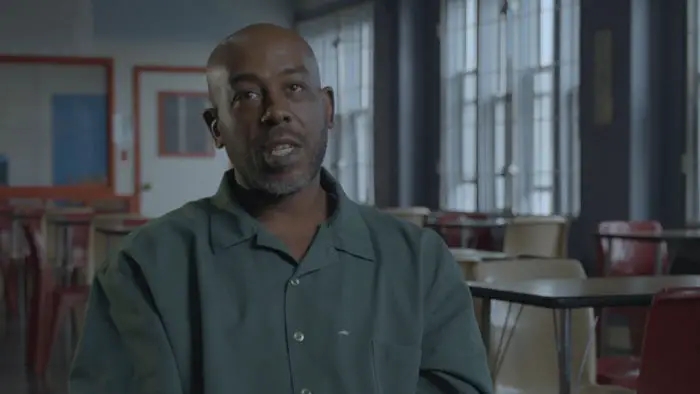 Interview shot of an incarcerated man in a green uniform speaking in a prison room with windows behind him. 