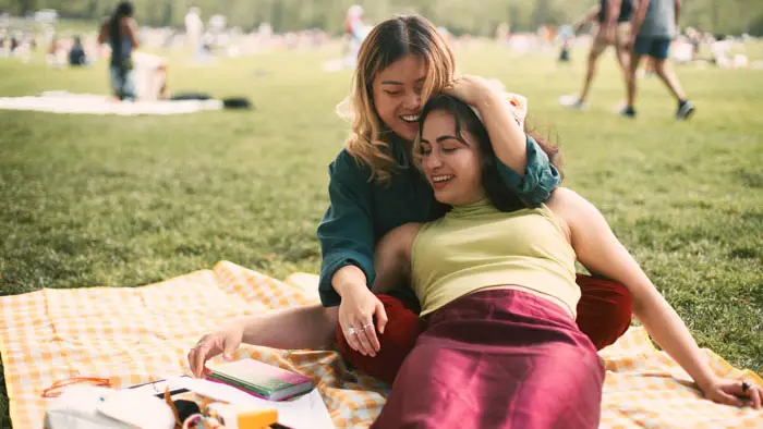 Two women relax on a picnic blanket in a park, smiling and leaning into each other.